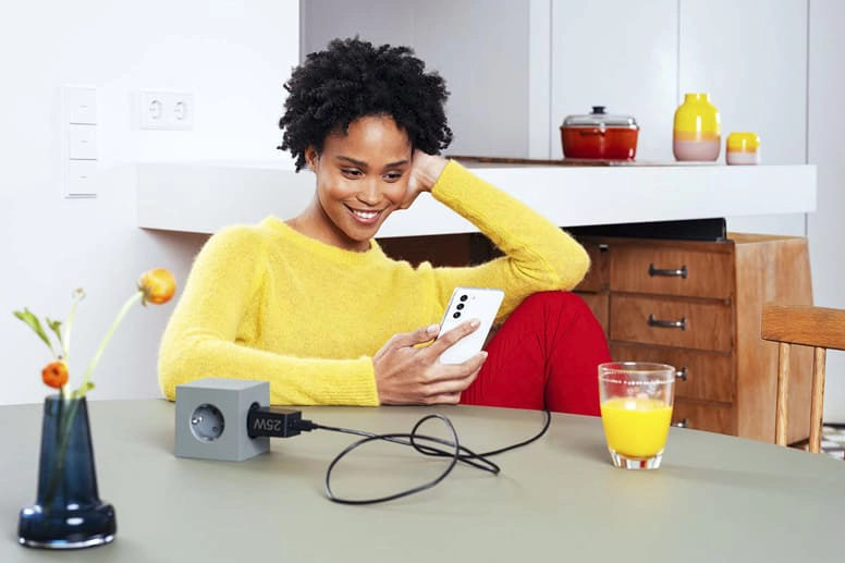 Una joven sentada a la mesa con un cargador rápido de Samsung sonríe a su smartphone.
