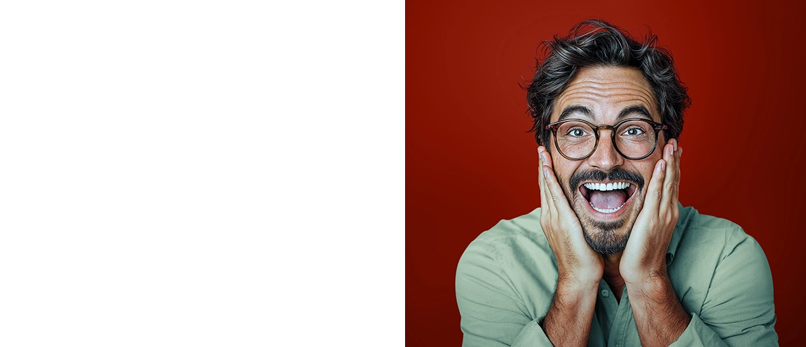 Un hombre de pelo negro y gafas oscuras mira a la cámara con la boca abierta por la emoción. Lleva un jersey verde claro y está de pie frente a una pared roja oscura.