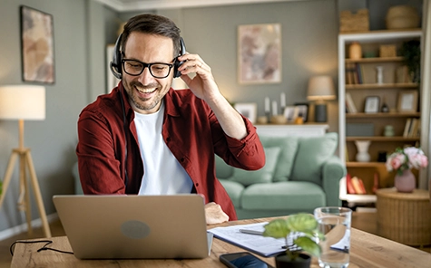 Un hombre trabaja desde casa, en el salón de su casa, con su ordenador portátil y habla por teléfono utilizando unos auriculares.