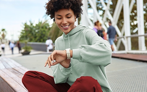 Una joven con jersey verde y pantalones rojos se sienta en una pared y mira su smartwatch de buen humor