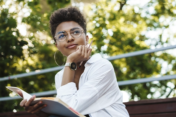 Una mujer con un look de negocios se sienta en un banco bajo los árboles y mira soñadoramente más allá de la cámara. Lleva el Hama Smartwatch «7000» en la muñeca.