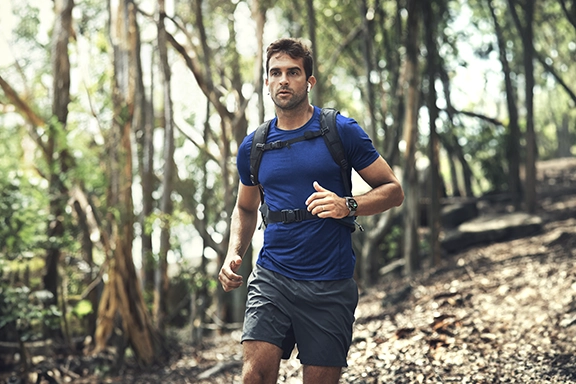 Un hombre con camiseta azul y pantalones cortos hace footing cuesta abajo por un bosque. Lleva el Hama Smartwatch '9000' en la muñeca.