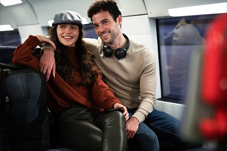 Una pareja está sentada en el tren. El hombre lleva unos auriculares al cuello y pasa el brazo por los hombros de su amiga.