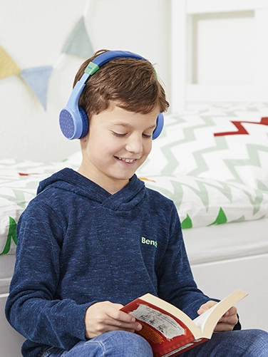 Un chico se sienta en su habitación y lee un libro mientras lleva puestos los auriculares Hama Bluetooth® "Teens Guard"