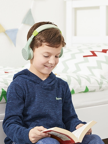 Un chico se sienta en su habitación y lee un libro mientras lleva puestos los auriculares Hama Bluetooth® "Teens Guard"