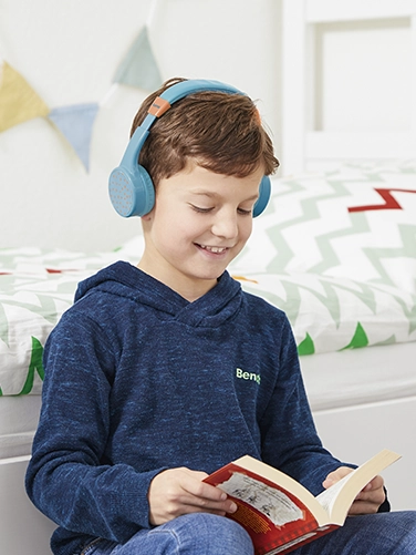 Un chico se sienta en su habitación y lee un libro mientras lleva puestos los auriculares Hama Bluetooth® «Teens Guard II»