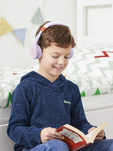 Un chico se sienta en su habitación y lee un libro mientras lleva puestos los auriculares Hama Bluetooth® «Teens Guard II»