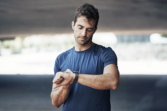 Un hombre con una camiseta azul se encuentra al aire libre bajo un puente en verano y mira su Hama Smartwatch «5000».