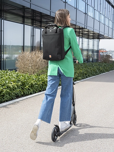 Una joven se aleja del espectador en un e-scooter con la mochila para portátil «Extreme Protect» de Hama a la espalda.