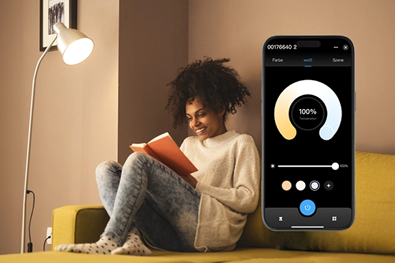 A la derecha, se ve un smartphone con la aplicación Smart Home abierta. A la izquierda y al fondo, una mujer se sienta relajada en el sofá y lee un libro bajo una lámpara de pie situada a su lado.