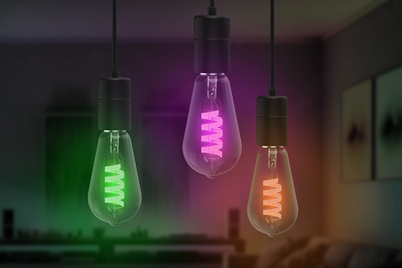 La elegante lámpara LED de Hama cuelga tres veces una al lado de la otra desde la parte superior del cuadro, en primer plano. Cada bombilla se ilumina de forma diferente: verde, rosa y naranja. Al fondo se ve un salón oscurecido y difuminado.<br>