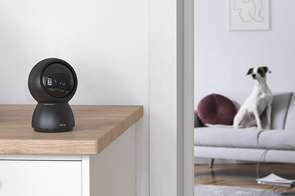 La cámara de vigilancia giratoria Hama con WLAN y detector de movimiento para interiores se encuentra en primer plano sobre un aparador de madera. Un perro está sentado en el sofá del fondo.